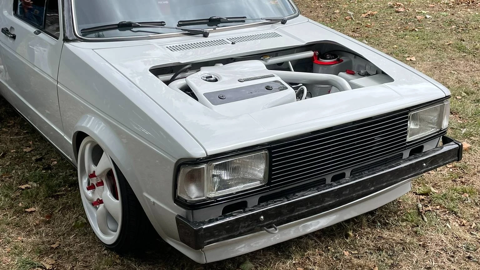 Front side view of a stanced bright gray Volkswagen Caddy. The cut hood reveals a swapped engine.