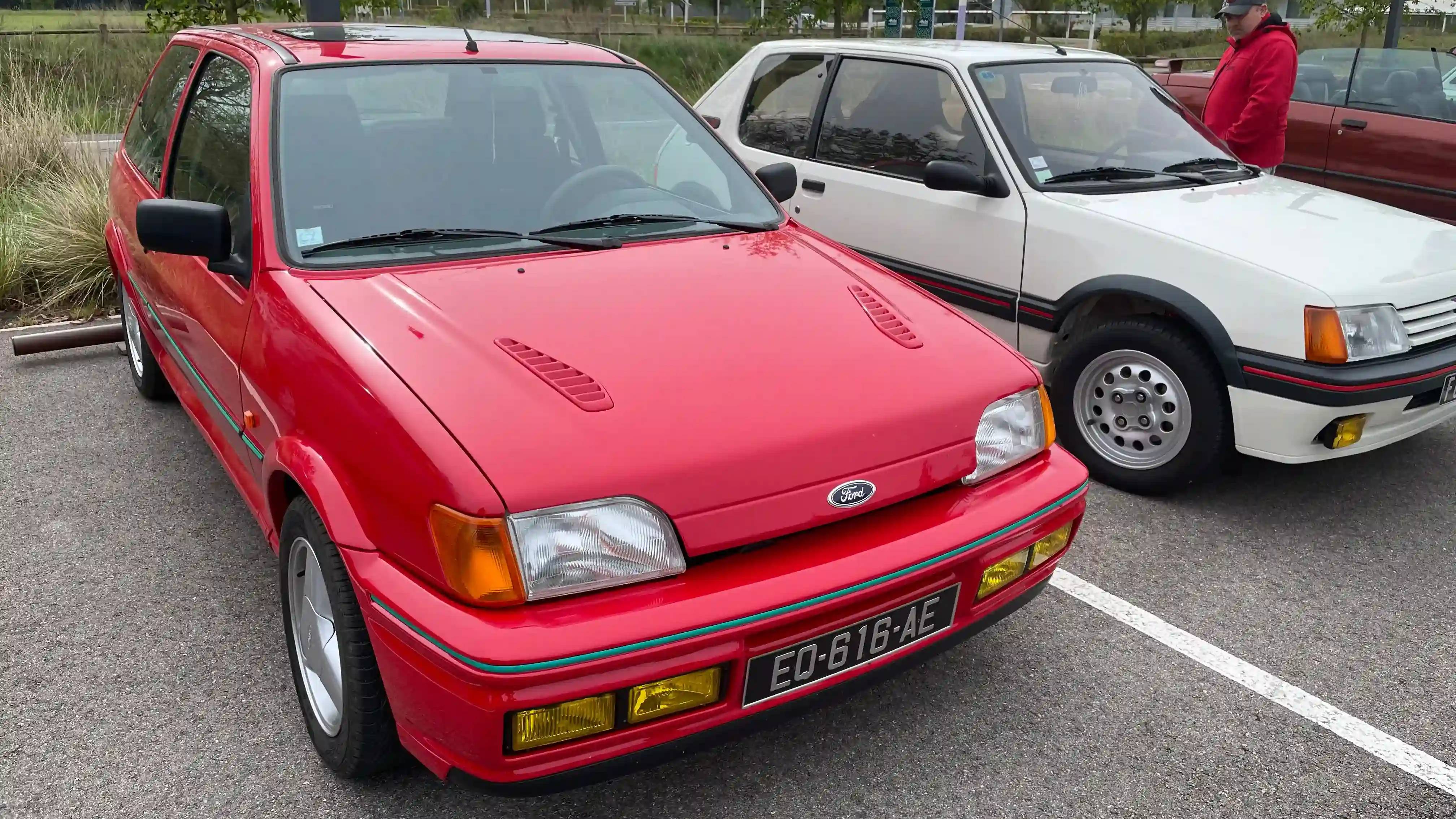 A red Ford Fiesta RS Turbo next to a white Peugeot 205 GTi, both parked