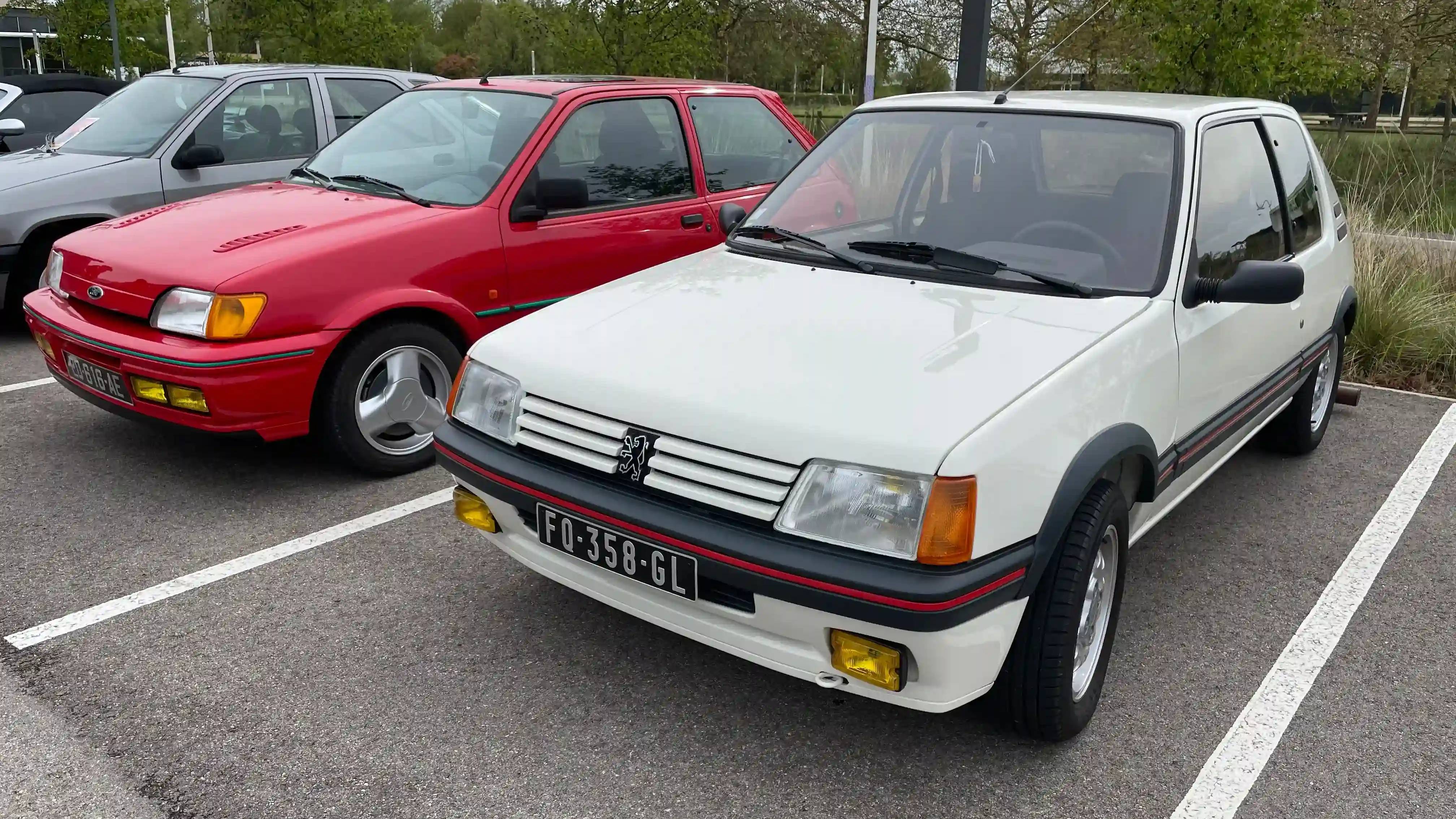 A red Ford Fiesta RS Turbo next to a white Peugeot 205 GTi, both parked