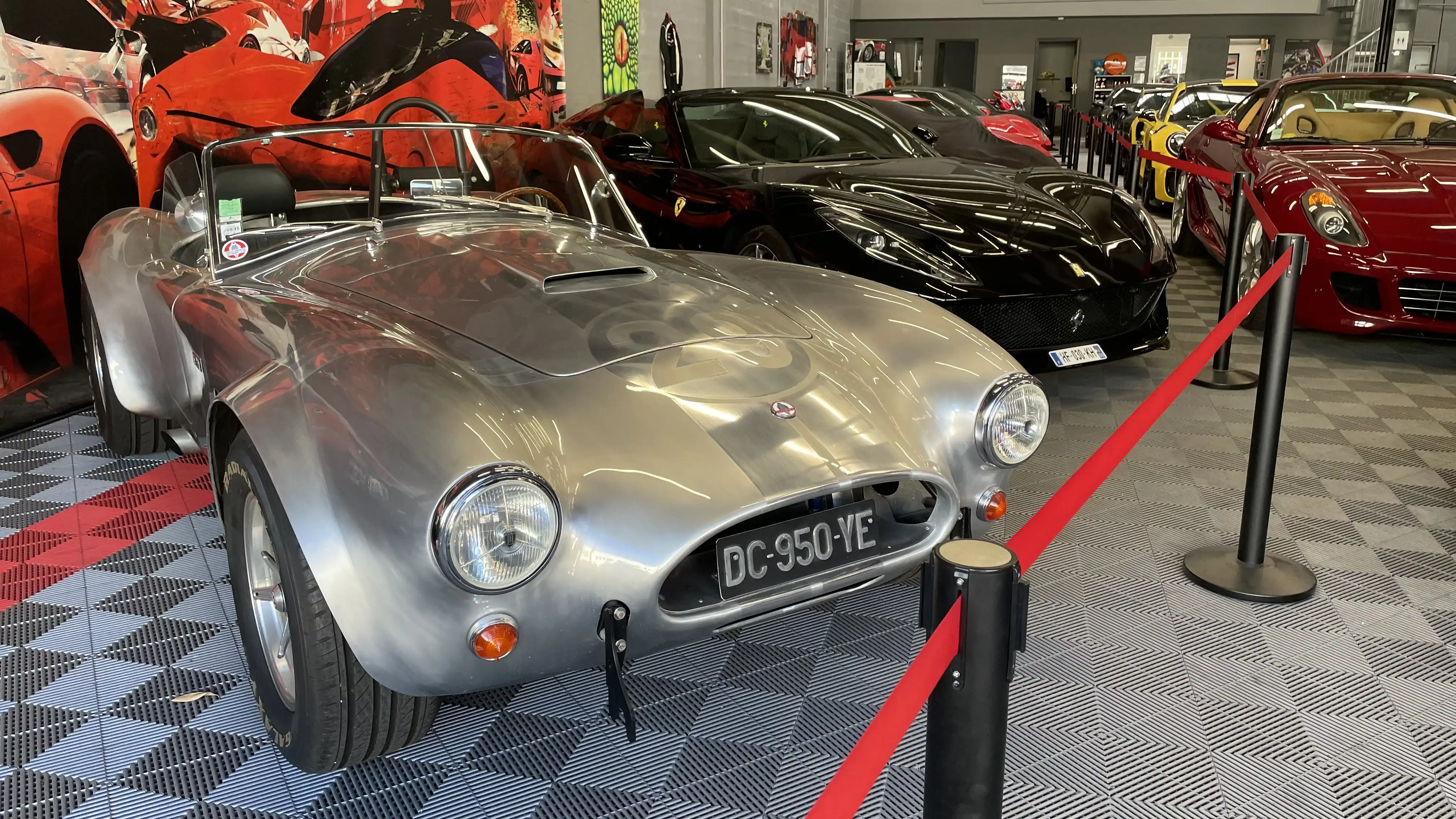 A grey Backdraft RT3 Cobra replica in front of various luxury cars inside the showroom of Dream Car Performance